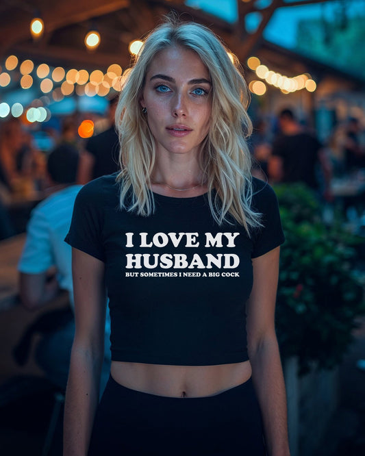 Blonde woman wearing a black crop top with bold white text that reads “I Love My Husband But Sometimes I Need a Big Cock” from Wicked Boutique®