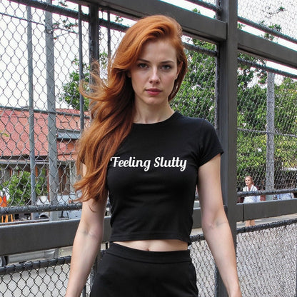 Redhead woman wearing a black crop top with “Feeling Slutty” in white text. Flirty kink shirt for bratty submissives and public play teasing.