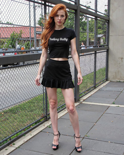 Redhead woman wearing a black crop top with “Feeling Slutty” in white text. Flirty kink shirt for bratty submissives and public play teasing.