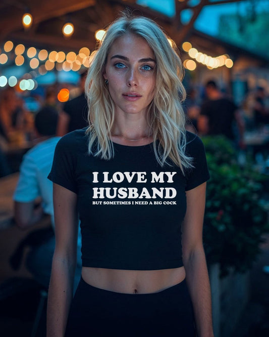 Blonde woman wearing a black crop top with bold white text that reads “I Love My Husband But Sometimes I Need a Big Cock” from Wicked Boutique®