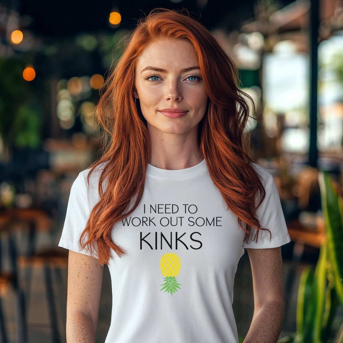 Redhead wearing white unisex t-shirt with “I Need To Work Out Some Kinks” printed in bold black text and upside down pineapple graphic. Kinky workout shirt blending BDSM humor and fitness vibes.